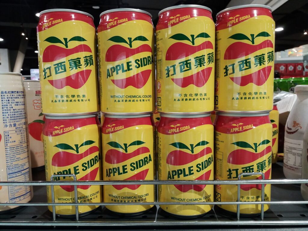 蘋果西打,Cans of Apple Sidra on a store shelf, featuring a yellow background, red apple design, and text highlighting "WITHOUT CHEMICAL COLORS."