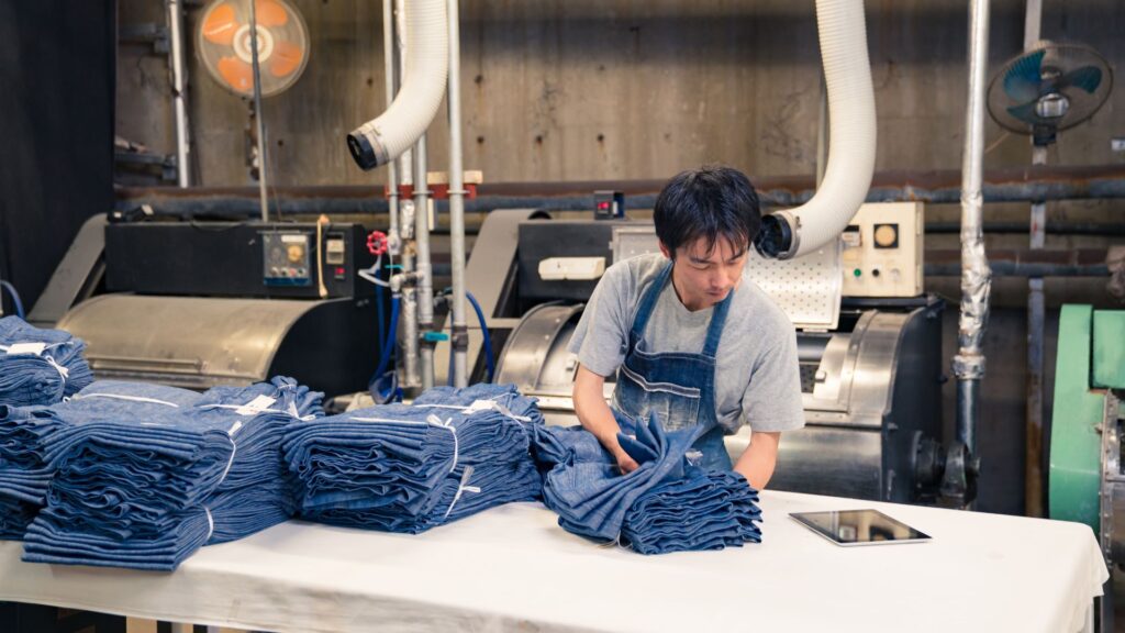 A worker wearing an apron is organizing neatly folded stacks of denim fabric on a table in an industrial setting, with machinery and ventilation pipes in the background. An electronic tablet lies on the table beside the fabrics.